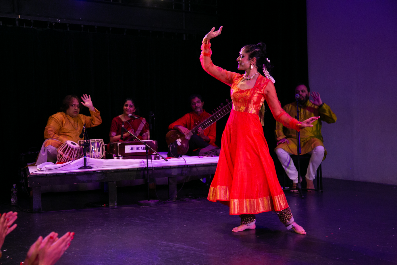 In front of the seated musicians, Rachna Nivas lifts one curved arm in the air. The other is curved to the side. Her body faces front, but her face is in profile.